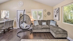 A living room with a sectional sofa, a hanging chair, a dining area with a table and chairs, and a large window with a view of greenery. - Bay Area Vacation Rental 4 Mi to Stanford! (Redwood City)