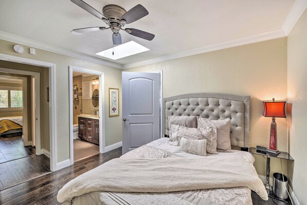 A bedroom with a large bed, a ceiling fan, a nightstand with a lamp, and a door leading to a bathroom.