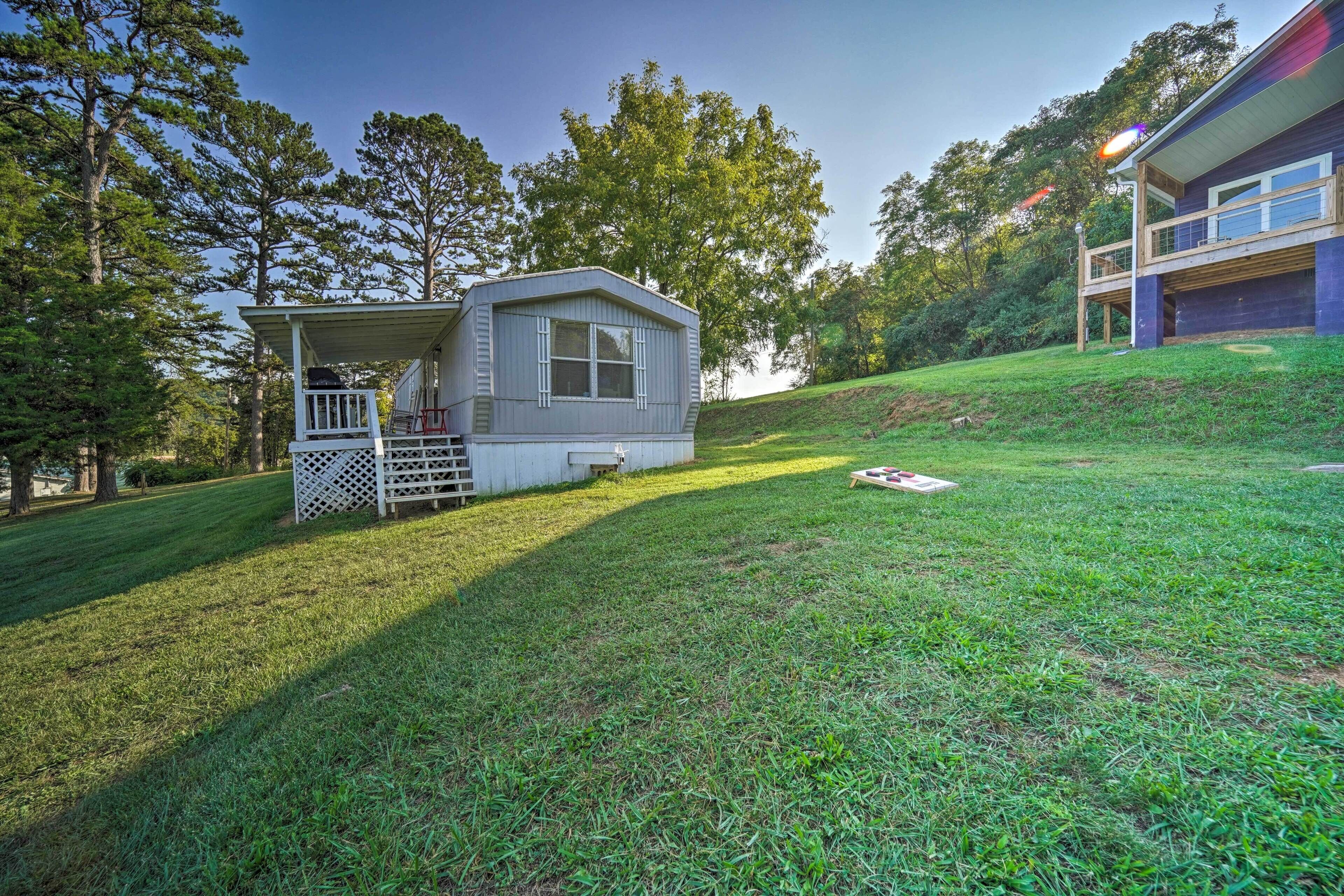 A small, single-story cabin with a porch and a deck, surrounded by a grassy yard and trees.