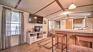 A cozy living area with a wooden floor, a TV mounted on the wall, a kitchen area with wooden cabinets, and a bar stool. - Quaint Cottage Overlooking Cherokee Reservoir (Bean Station)