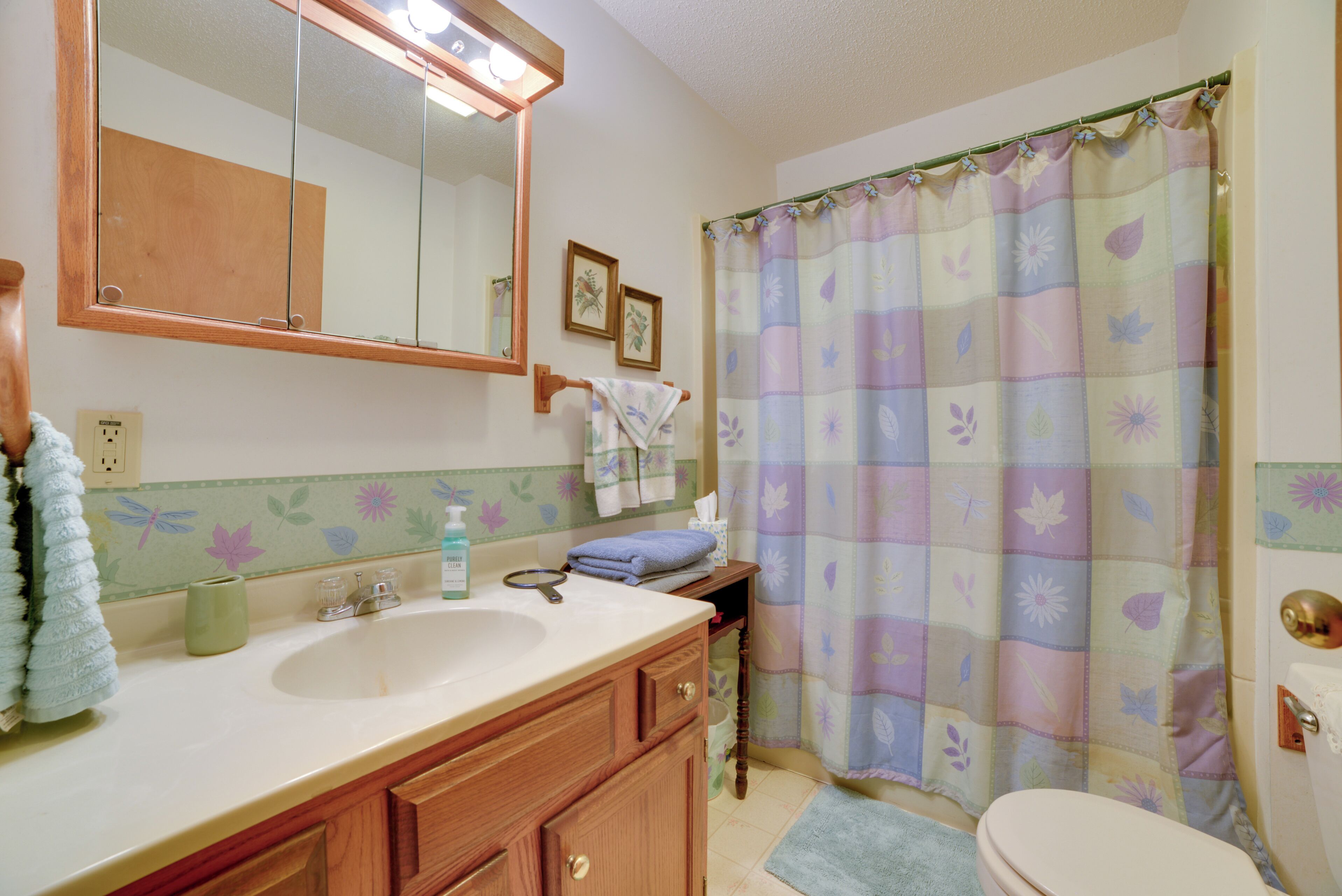 Une salle de bain avec un meuble-lavabo en bois, un grand lavabo, un miroir, un rideau de douche à motifs floraux et des toilettes.