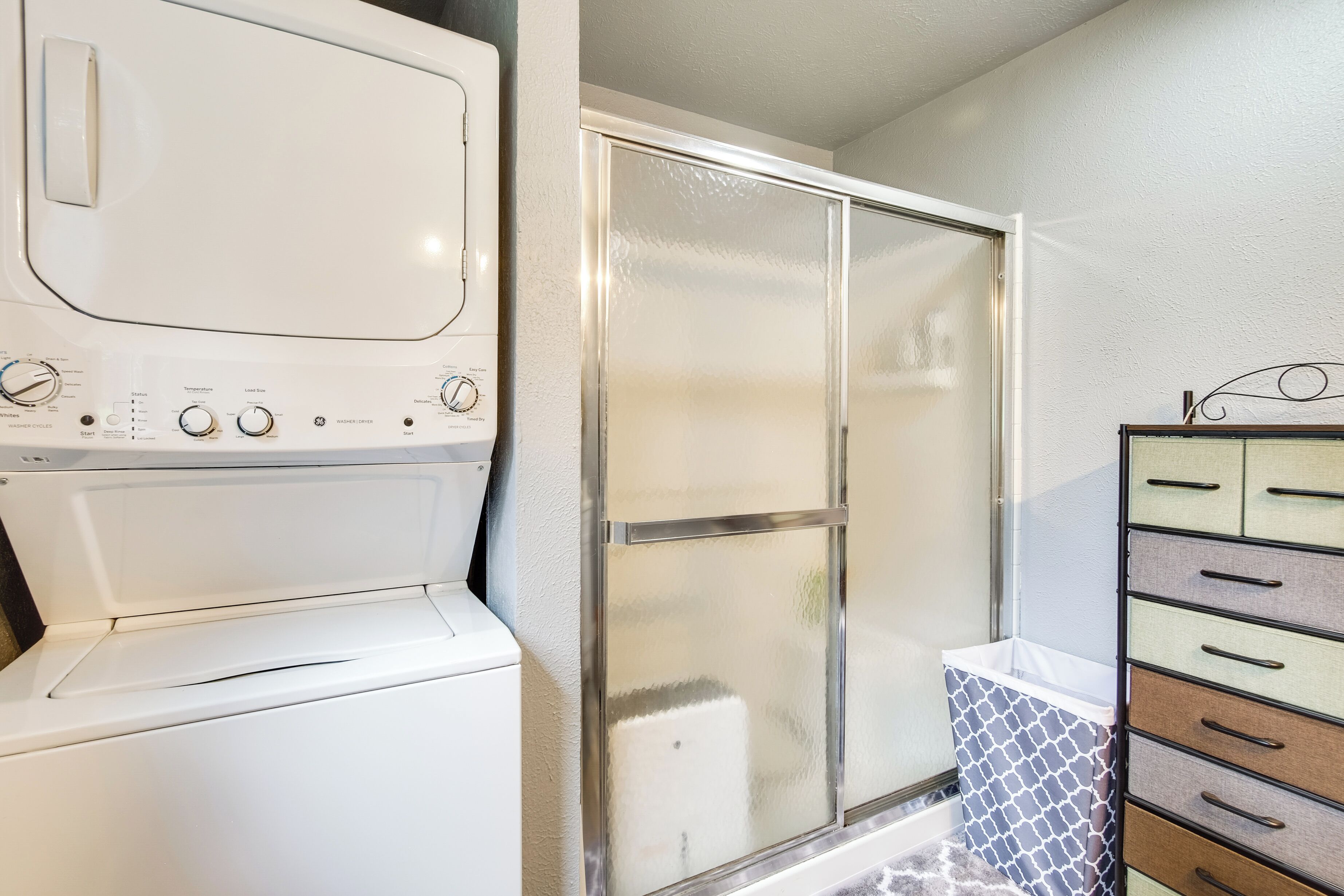 A laundry area with a front-loading washer and dryer, a glass shower enclosure, and a set of drawers with patterned storage.