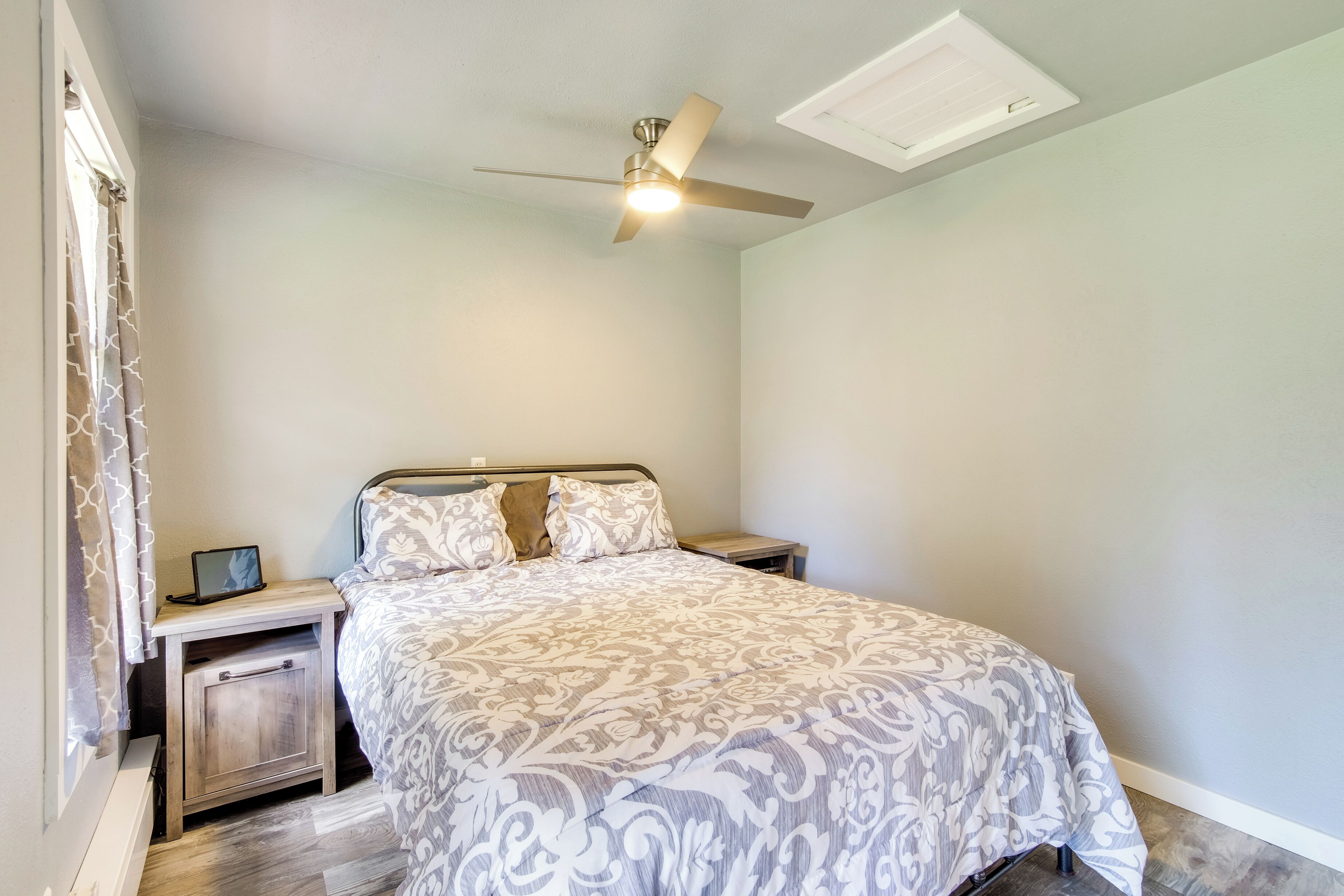 Un dormitorio con una cama, mesitas de noche, un ventilador de techo y una ventana con cortinas.