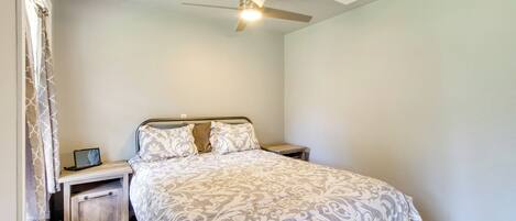 Une chambre à coucher comprenant un lit, des tables de chevet, un ventilateur de plafond et une fenêtre avec des rideaux.