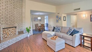 A living room with a brick fireplace, a grey sofa, a wooden coffee table, and a dining area in the background.
