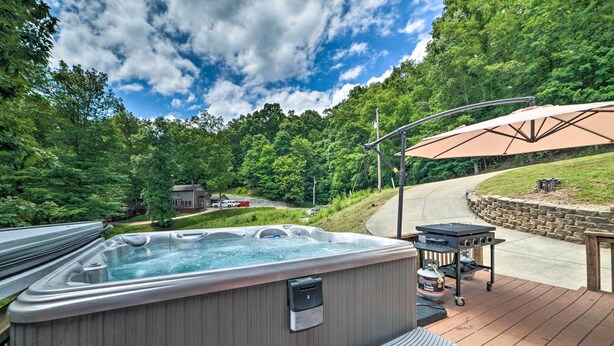 A hot tub on a wooden deck with a barbecue grill and umbrella, surrounded by lush greenery. - Lakefront 'rest-ashored' Home w/ Private Dock! (Eddyville)
