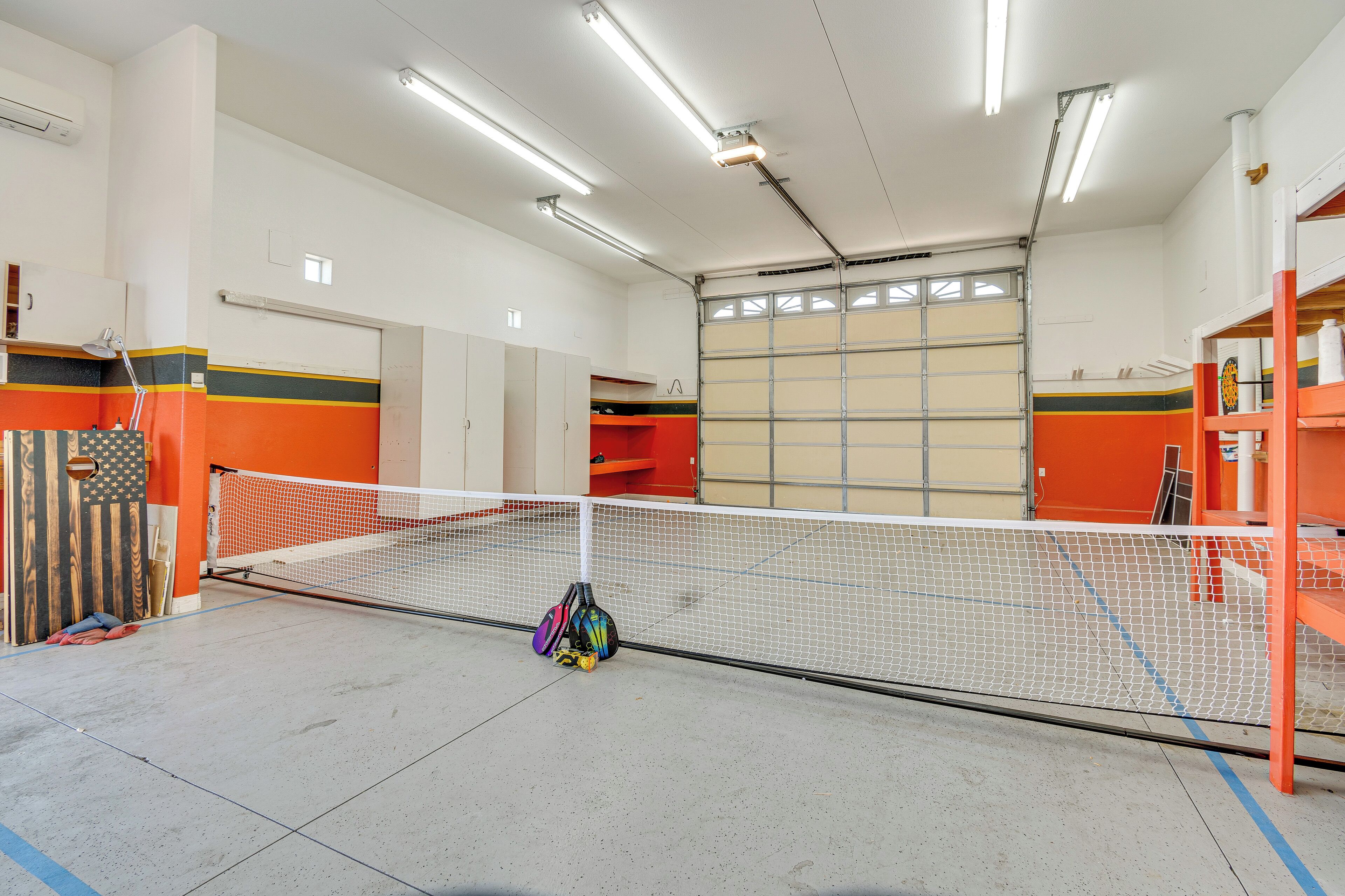 A tennis court with a net, surrounded by orange and beige walls, and equipped with overhead lighting.