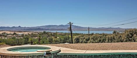 A pool with a curved design, surrounded by a pebble beach and a view of distant mountains and a body of water.
