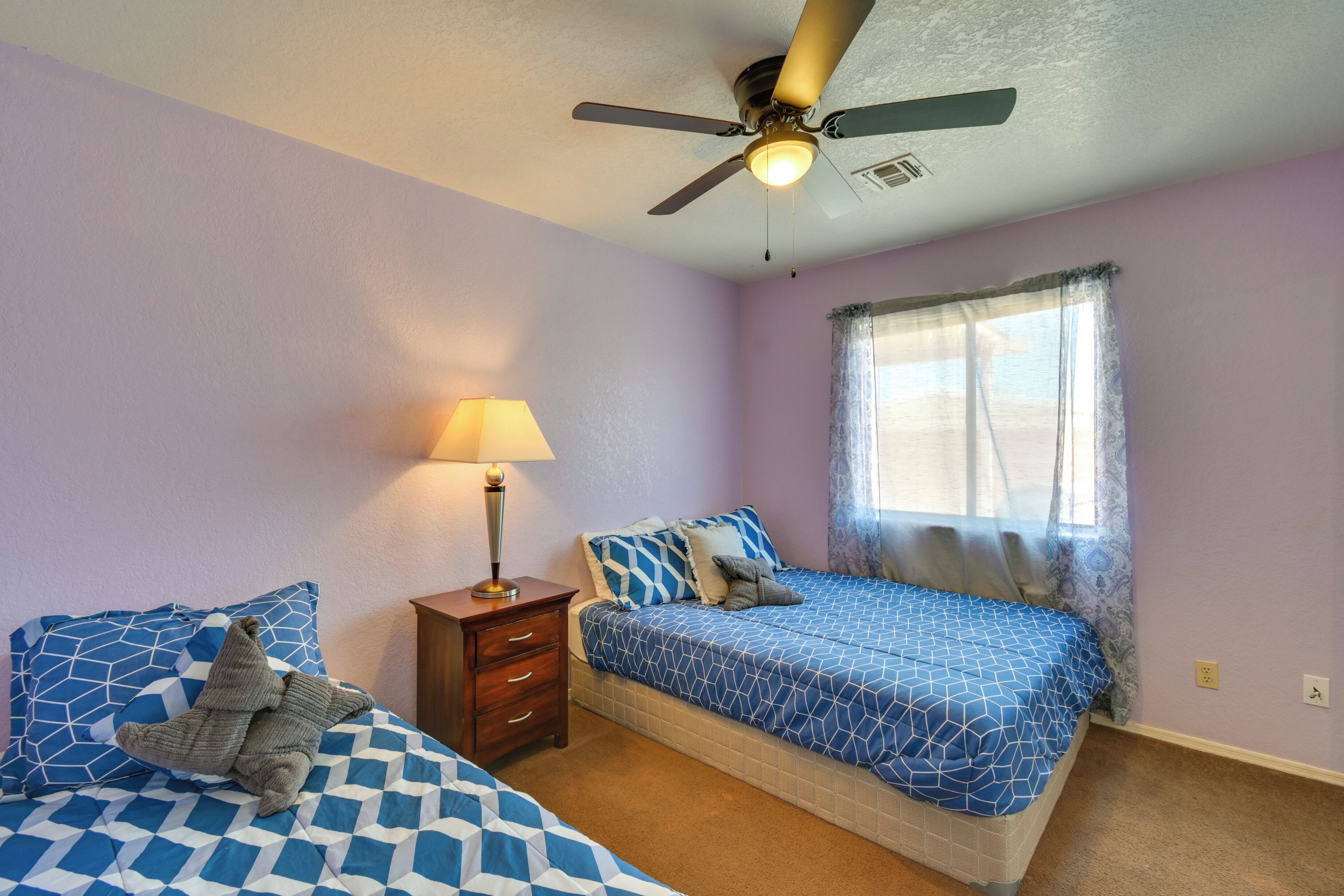 A bedroom with two beds, a ceiling fan, a nightstand with a lamp, and a window with sheer curtains.