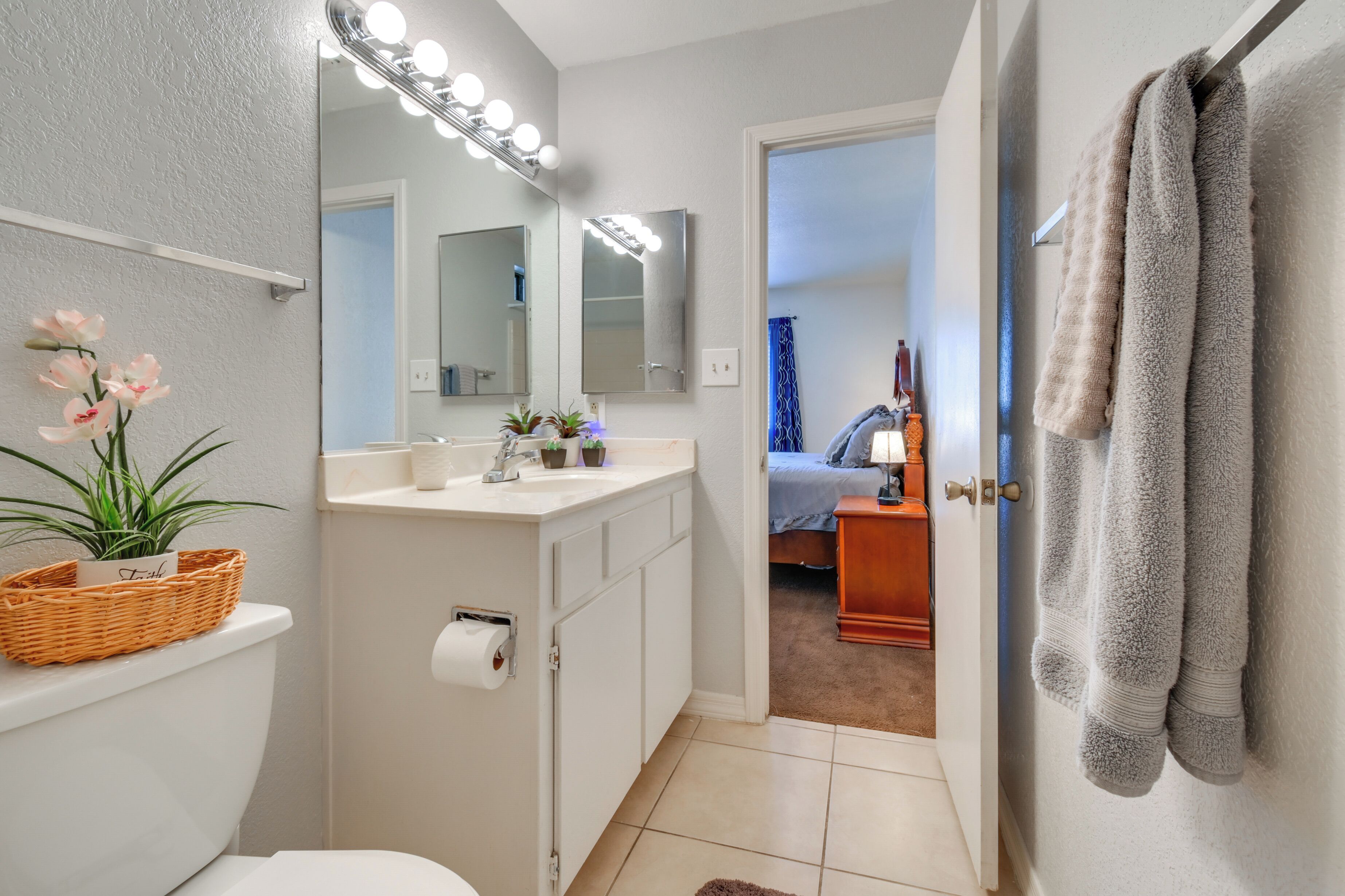 A bathroom with a toilet, sink, and mirror with lights. There is a basket on the toilet, a plant, and a towel rack.