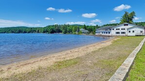 Une propriété au bord du lac, dotée d’une plage de sable, d’un grand bâtiment blanc avec un porche et, en arrière-plan, d’une colline boisée.