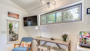 A living room with a rocking chair, a wooden table with stools, a chandelier, and a large window with a view of trees.