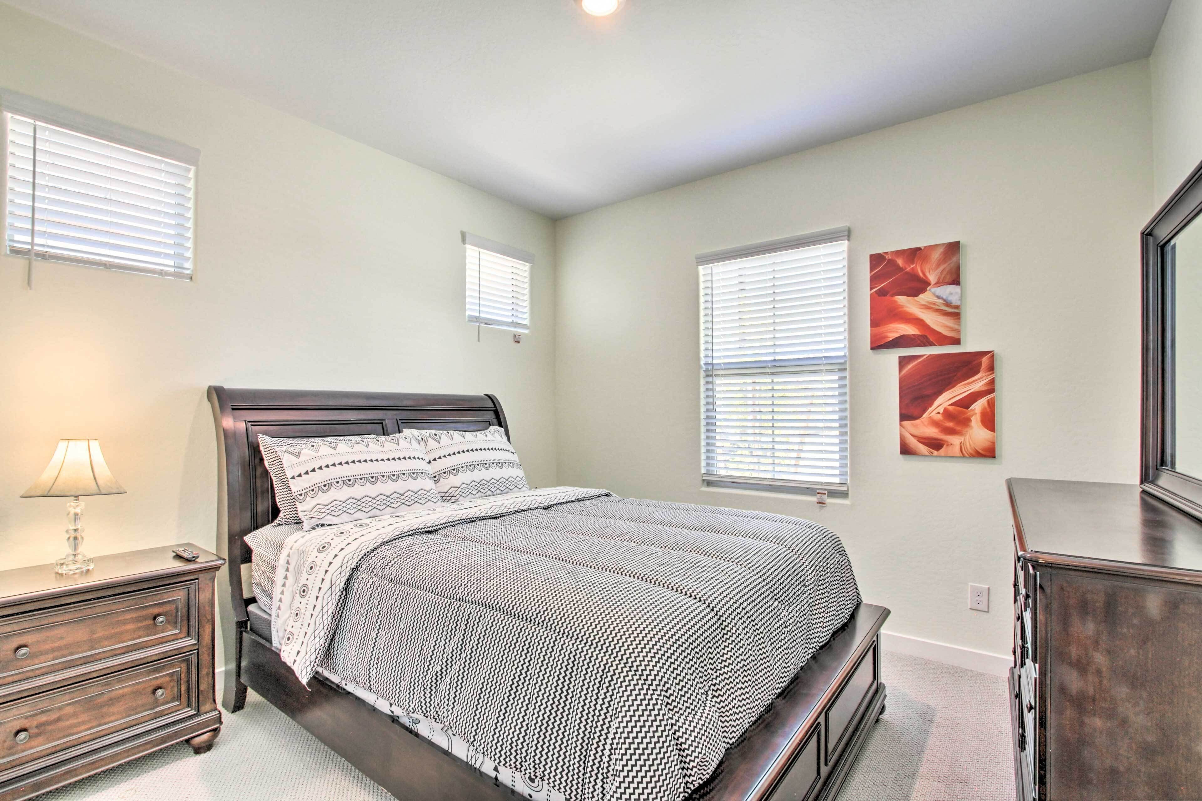 A bedroom with a bed, nightstand, dresser, and two windows with blinds.