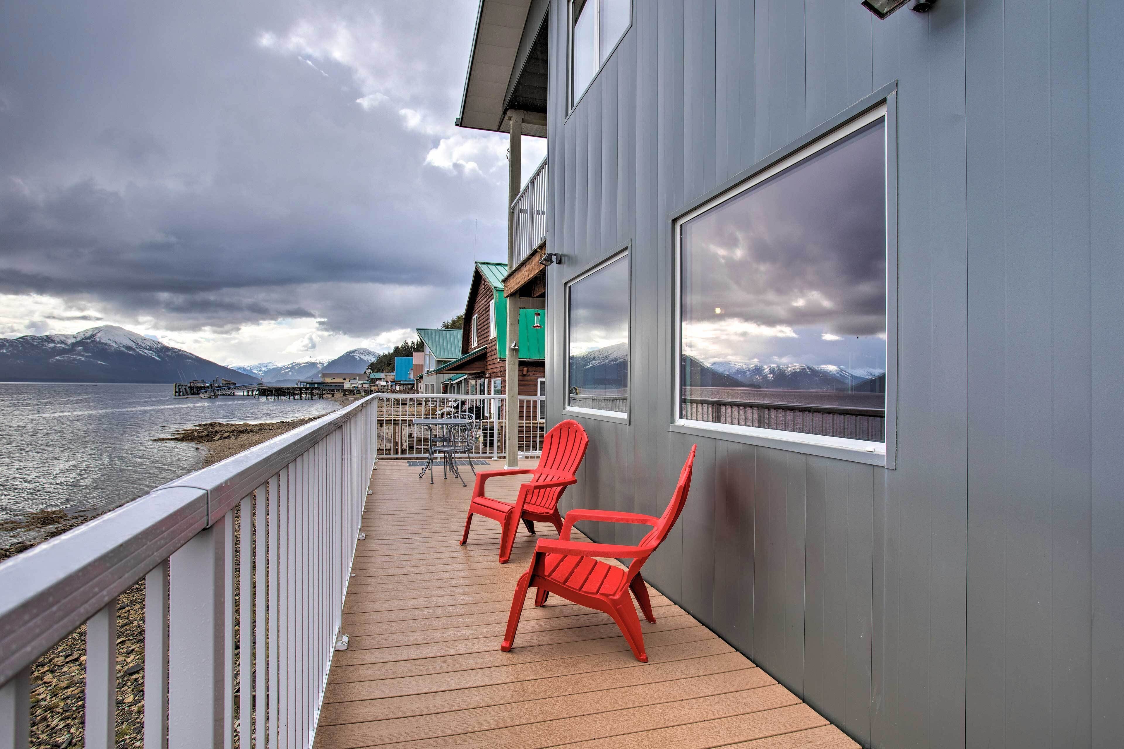 Une terrasse en bois avec deux fauteuils Adirondack rouges, un bâtiment gris doté de grandes fenêtres, et une vue sur des montagnes et une jetée en arrière-plan.