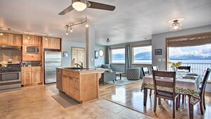 A modern kitchen with wooden cabinets, stainless steel appliances, and a large island. There is a dining area with a table and chairs, and a view of mountains through large windows.