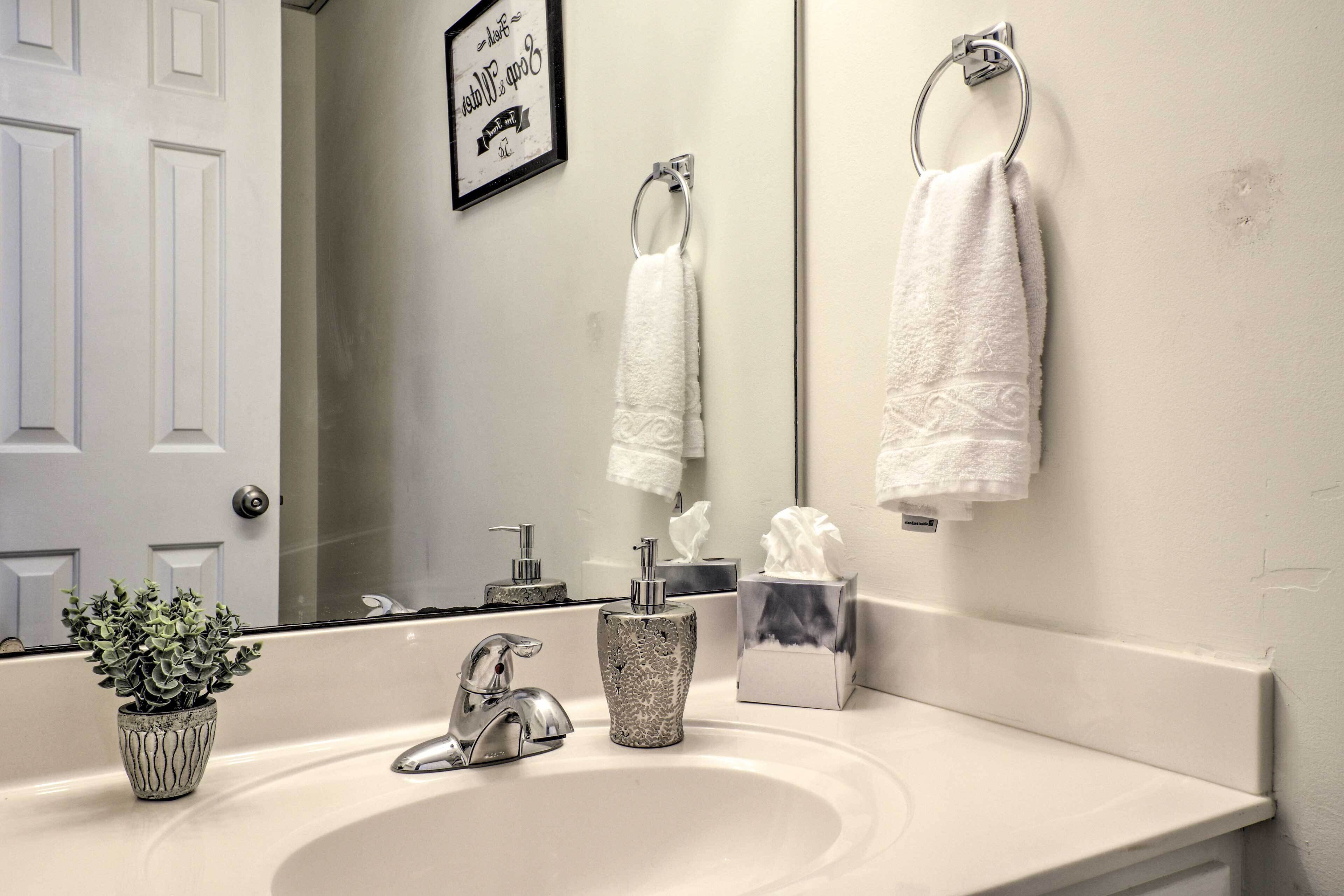 Une salle de bain avec un lavabo blanc, un miroir, un porte-serviettes, une plante en pot et un panneau encadré accroché au mur.