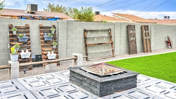 A backyard with a fire pit, potted plants, and a wooden shelf with hanging lights.