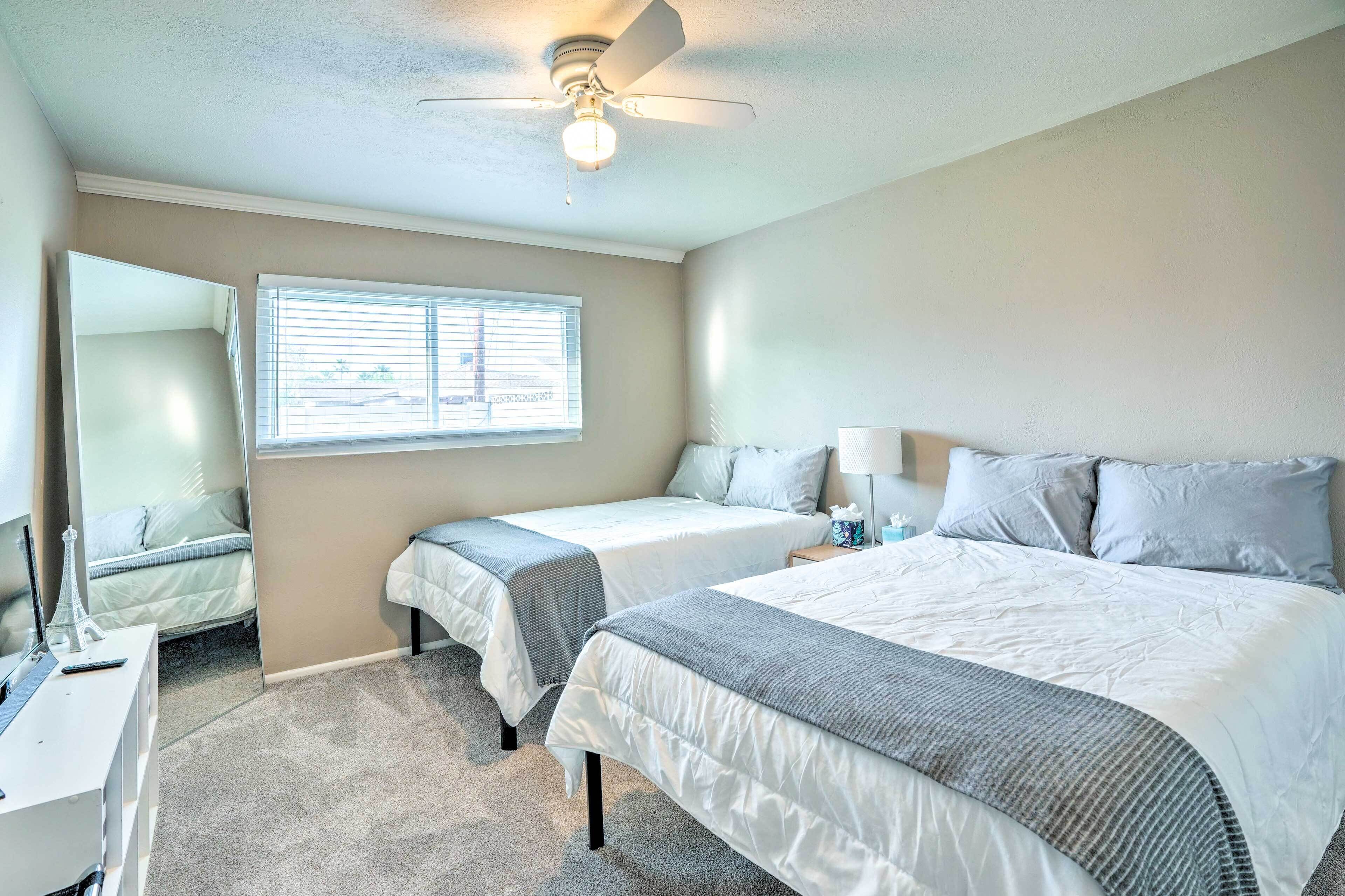 A hotel room with two beds, a ceiling fan, a window with blinds, a bedside table with a lamp, and a small desk with a lamp.