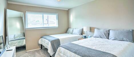 A hotel room with two beds, a ceiling fan, a window with blinds, a bedside table with a lamp, and a small desk with a lamp.