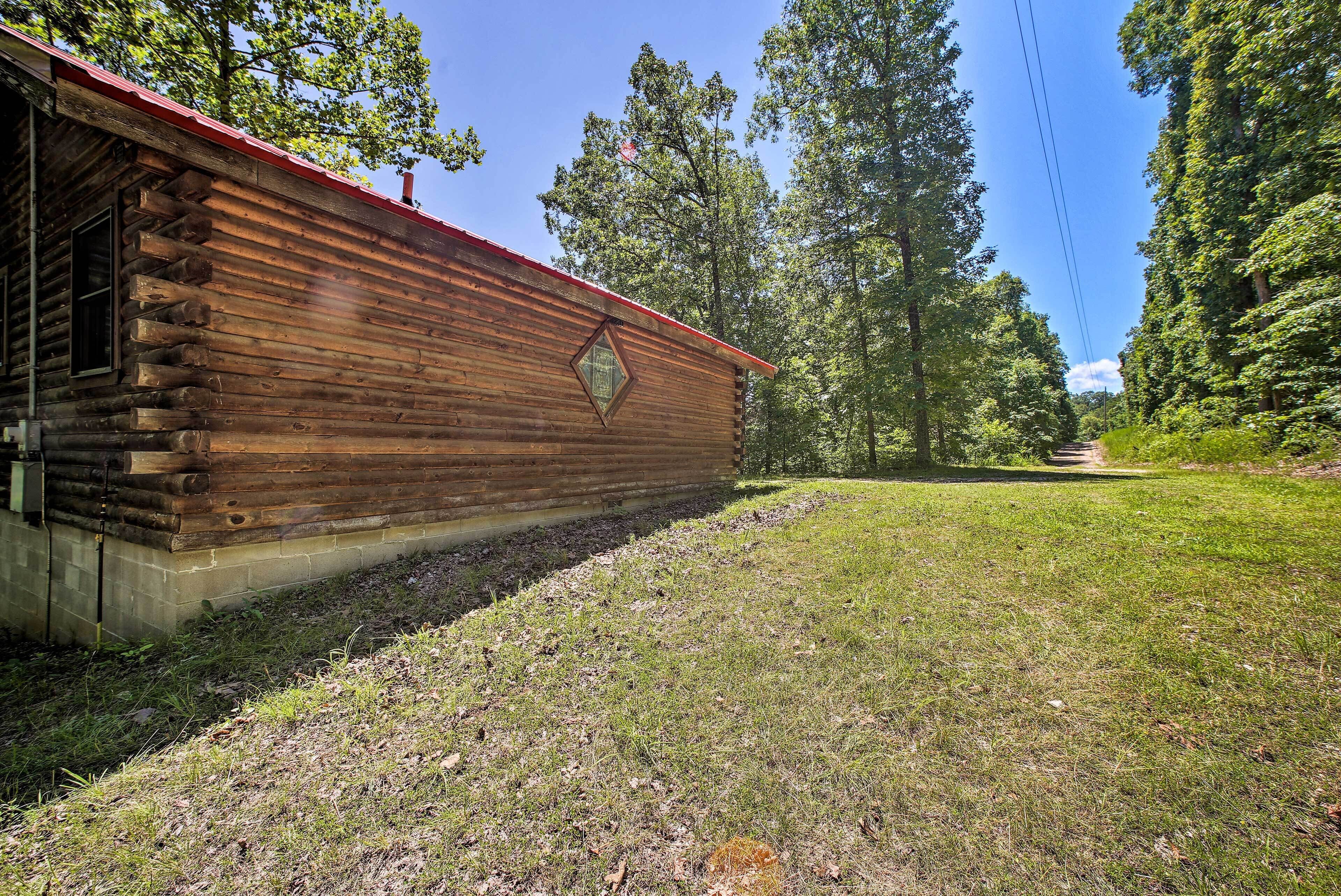 A rustic log cabin with a red roof, surrounded by a grassy area and tall trees.