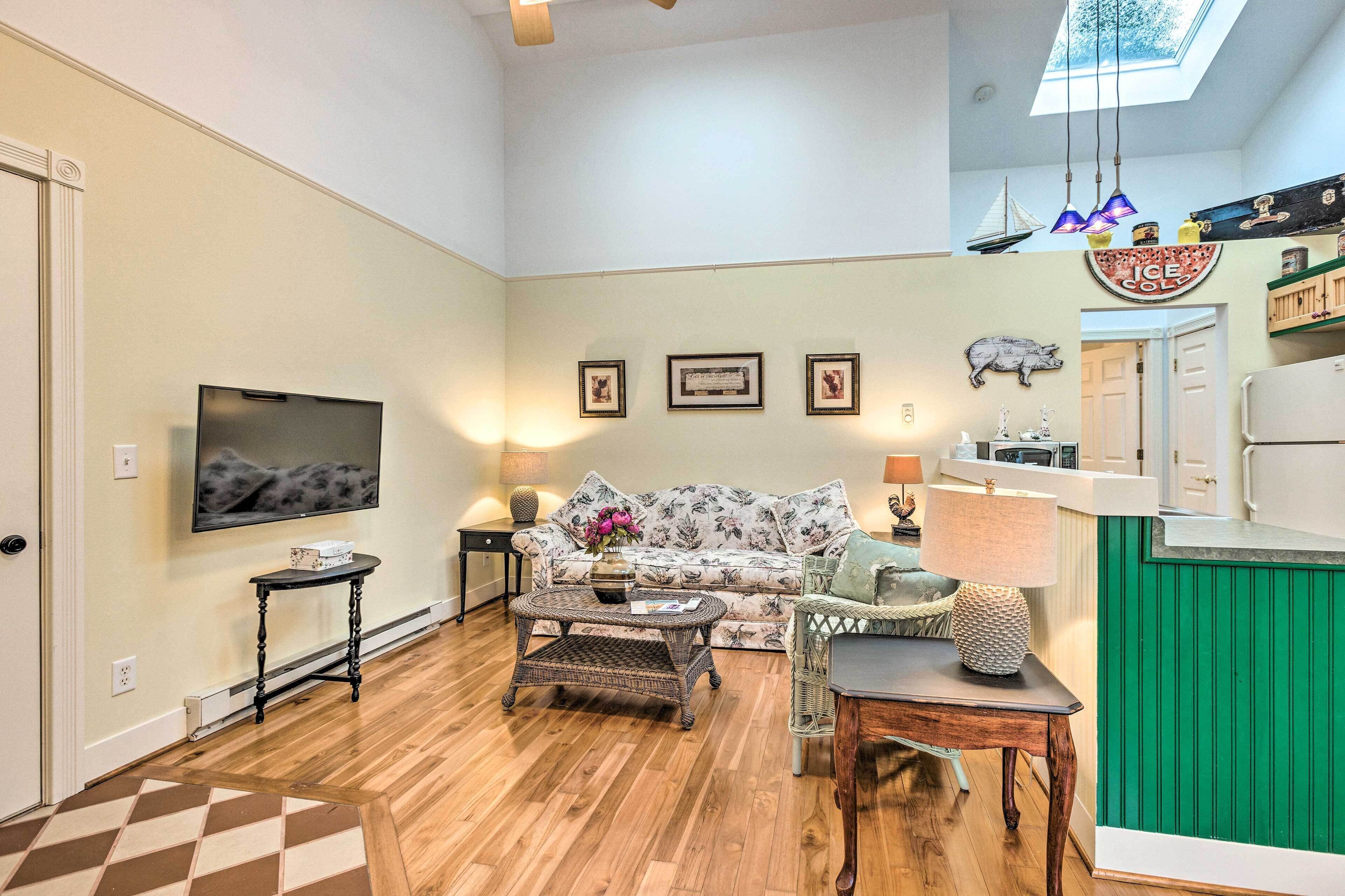 A spacious living room with a large flat-screen TV, a floral sofa, a coffee table, and a dining area with a green accent wall.
