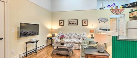 A spacious living room with a large flat-screen TV, a floral sofa, a coffee table, and a dining area with a green accent wall.