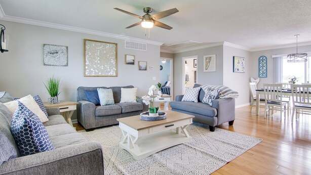 A spacious living room with a ceiling fan, grey sofas, a coffee table, and a dining area with a table and chairs. - Sunsets, Salt Air and Single-story Ease in Fenwick (Selbyville)