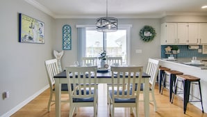 A dining area with a table set for four, a kitchen with white cabinets, and a view of the outdoors through a large window. - Sunsets, Salt Air and Single-story Ease in Fenwick (Selbyville)
