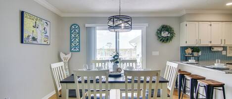 A dining area with a table set for four, a kitchen with white cabinets, and a view of the outdoors through a large window.