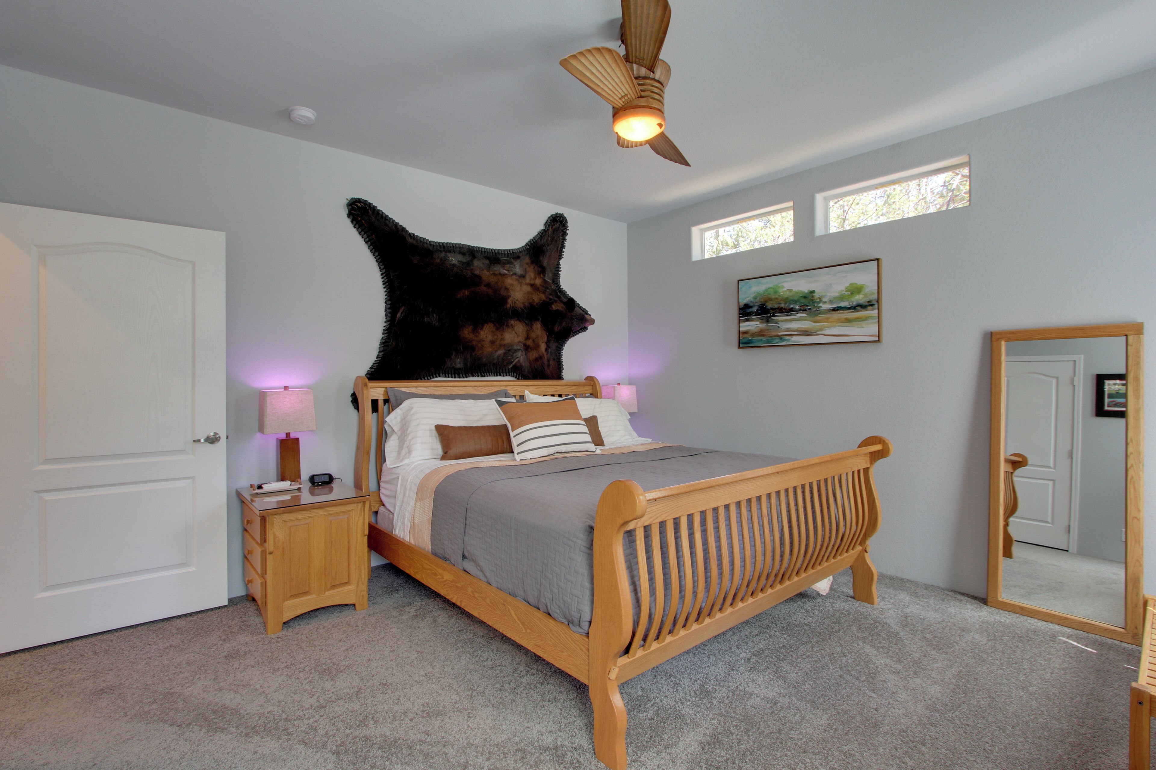 Une chambre avec un lit en bois, une table de chevet, une sculpture représentant une tête d’animal fixée au mur, un ventilateur de plafond et une fenêtre.