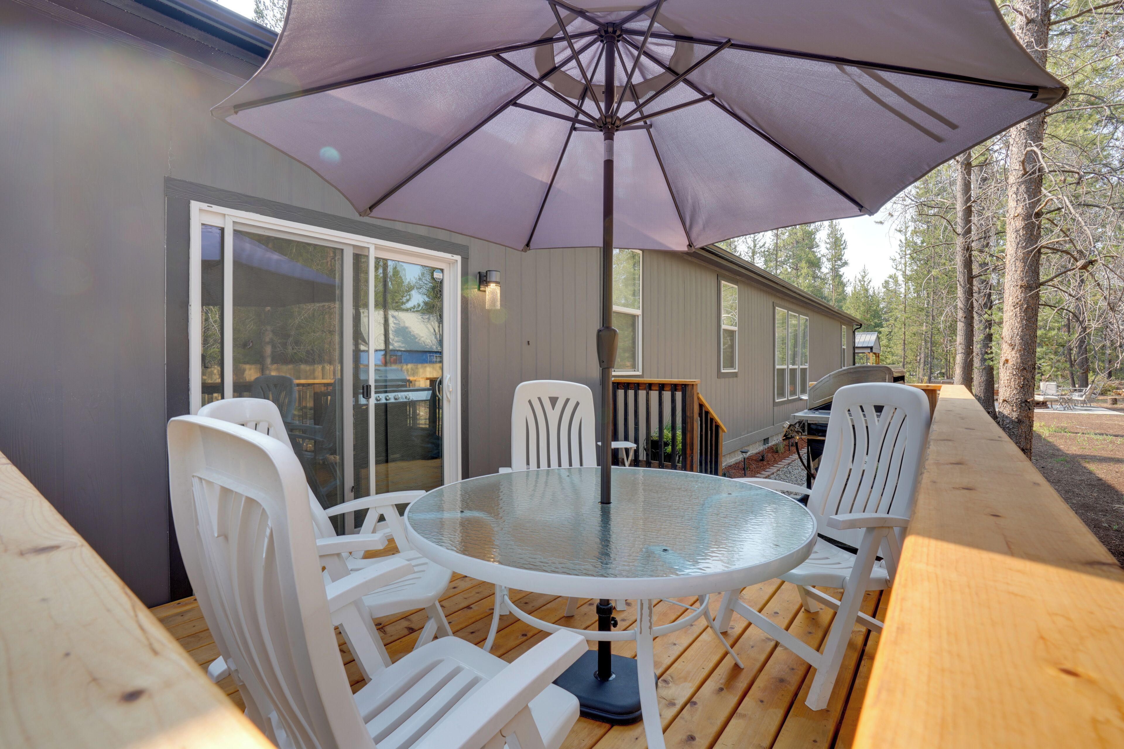 A wooden deck with a glass table and white chairs, an umbrella, and a house in the background.