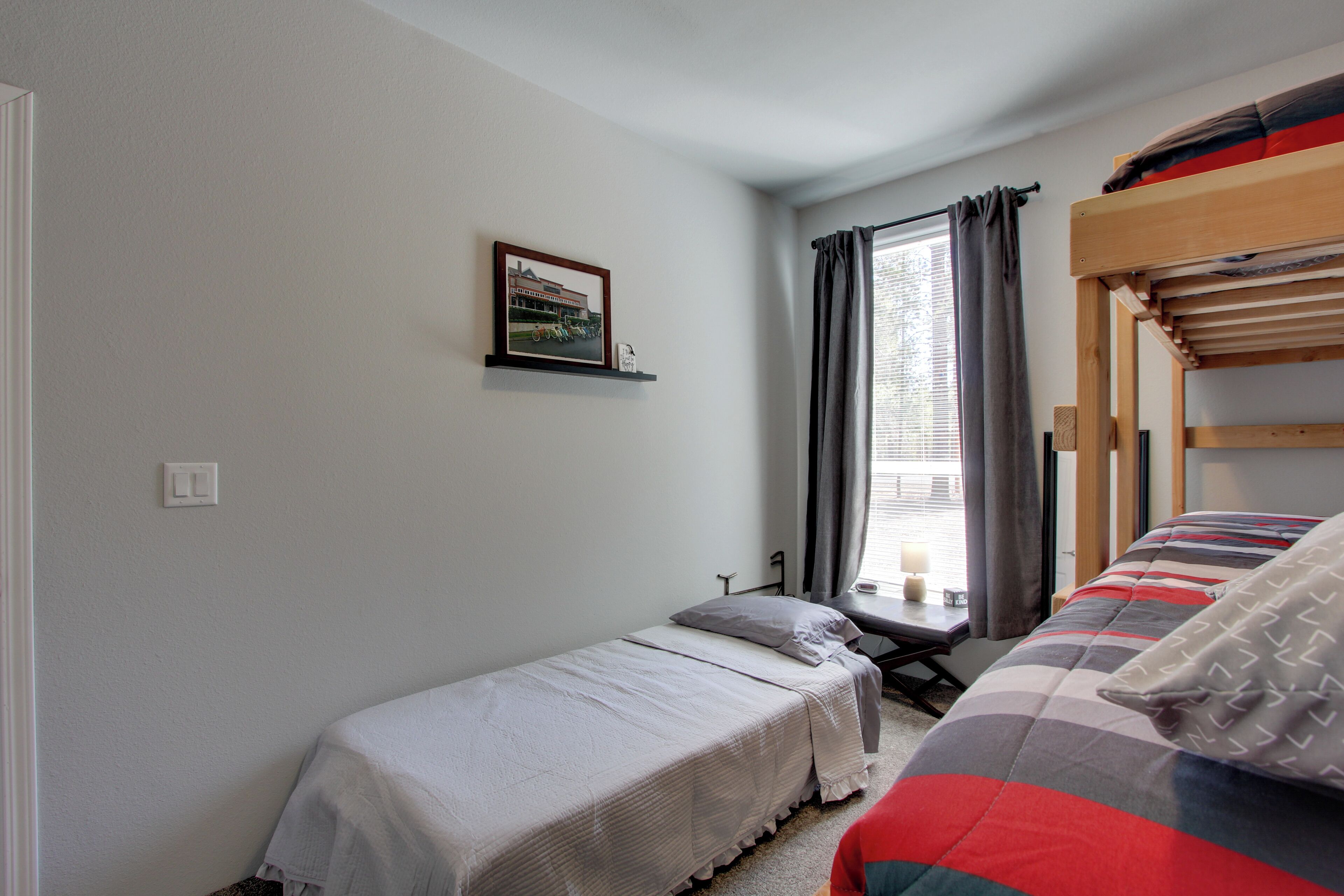 A bedroom with two single beds, a bunk bed, a window with curtains, a bedside table, and a framed picture on the wall.