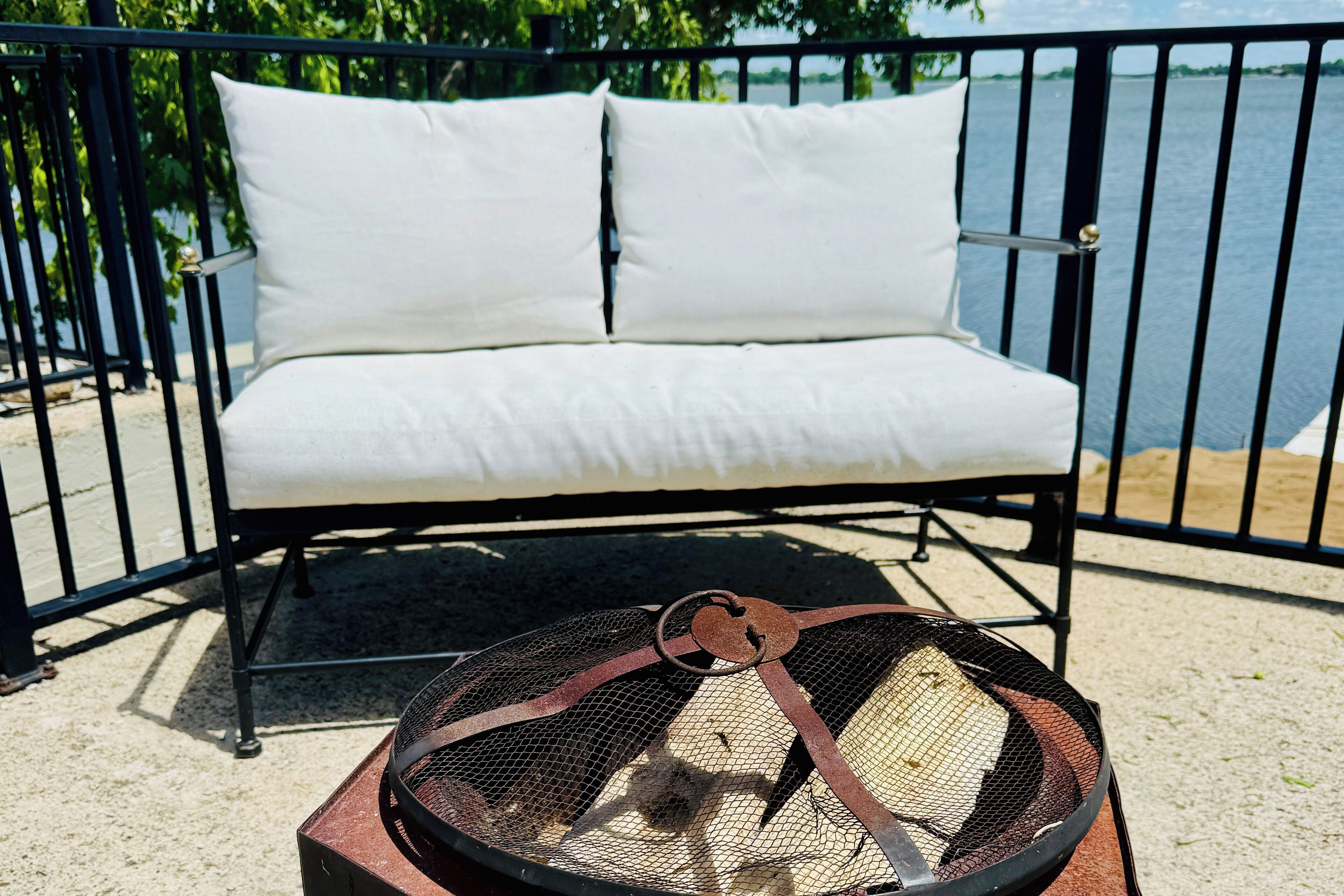 A black metal bench with white cushions, a metal fire pit with a mesh cover, and a body of water in the background.