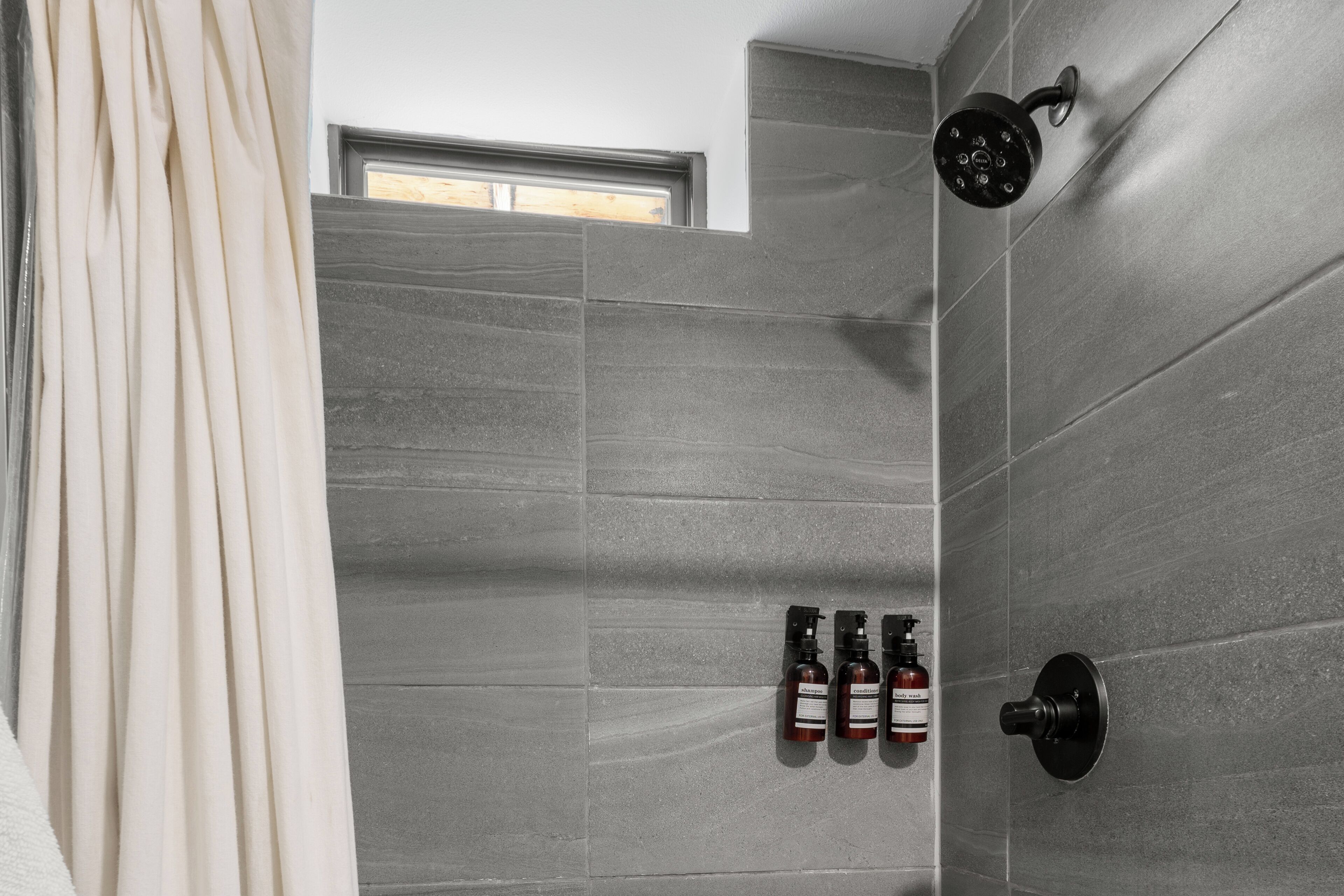 Un baño moderno con azulejos grises, un cabezal de ducha y tres botellas de artículos de aseo.