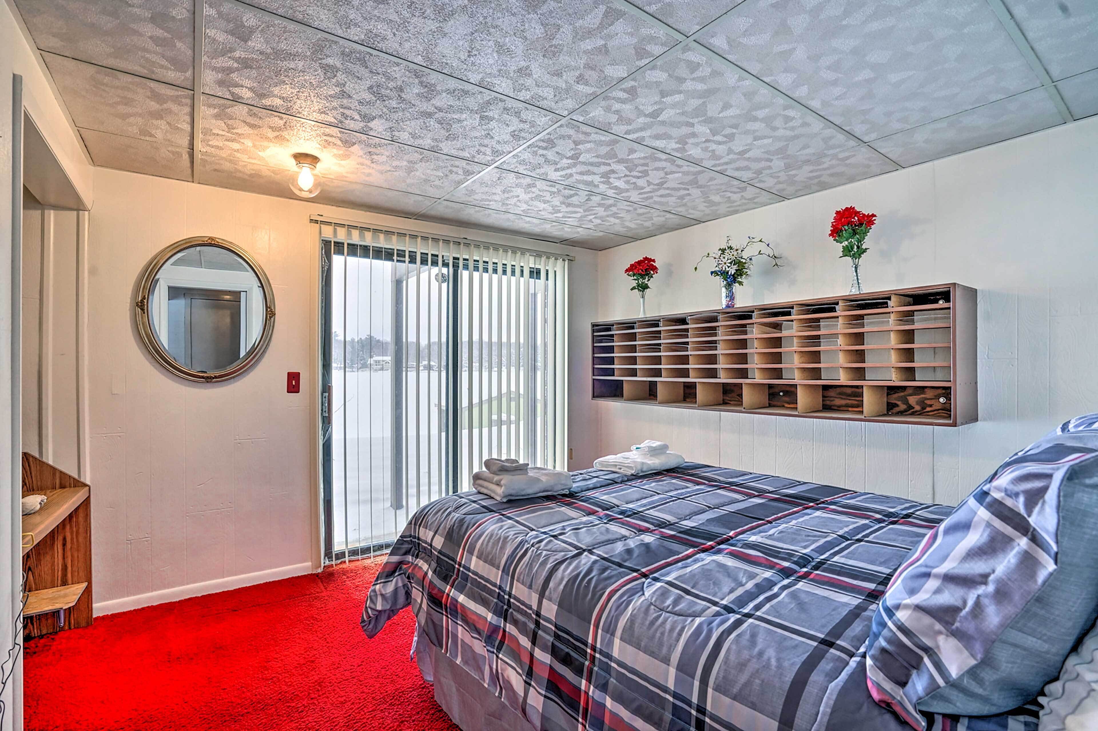 A hotel room with a red carpet, a bed with a plaid bedspread, a round mirror, a shelf with decorative items, and a view through the window.