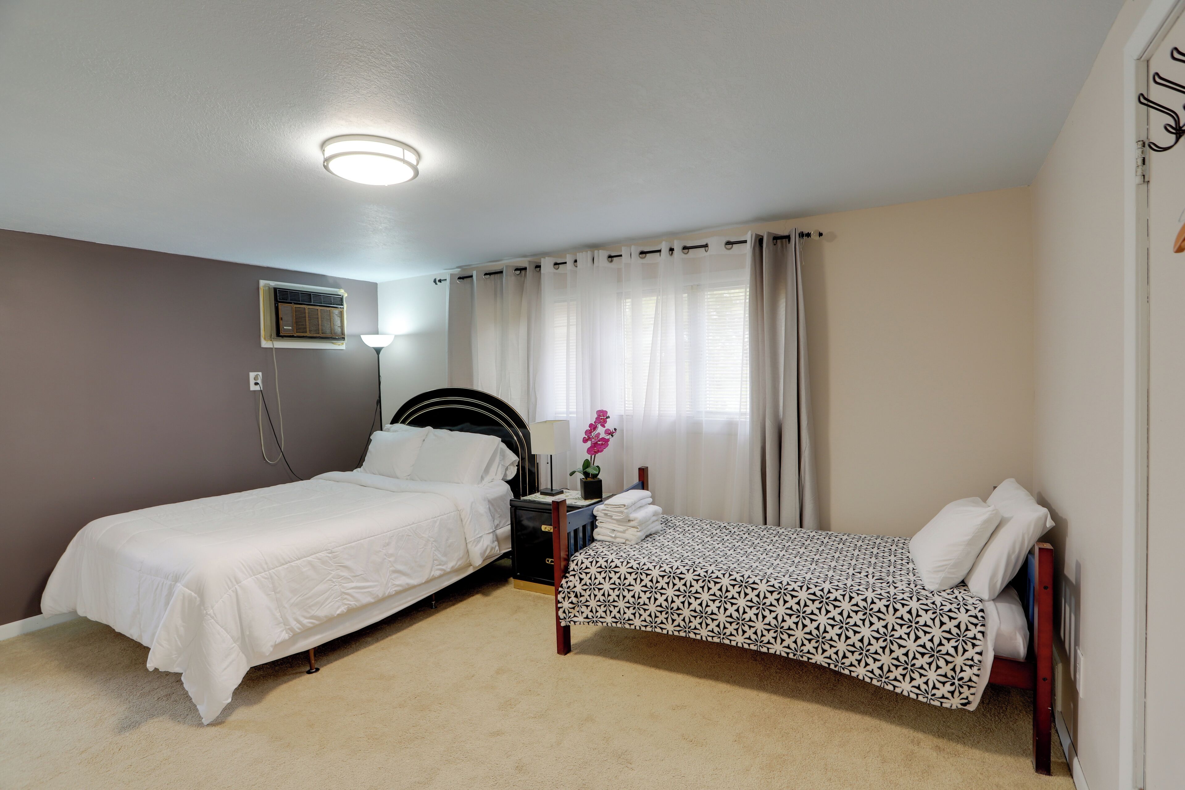A bedroom with two beds, a bedside table with a lamp, an air conditioning unit, and a window with curtains.