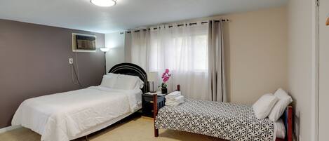 A bedroom with two beds, a bedside table with a lamp, an air conditioning unit, and a window with curtains.