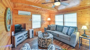 A cozy cabin interior with a wooden ceiling, a ceiling fan, a television, a sofa, a patterned ottoman, and a small table with a lamp. - Our Lakeside Retreat w/ Deck on Lake Herrington! (Lancaster)