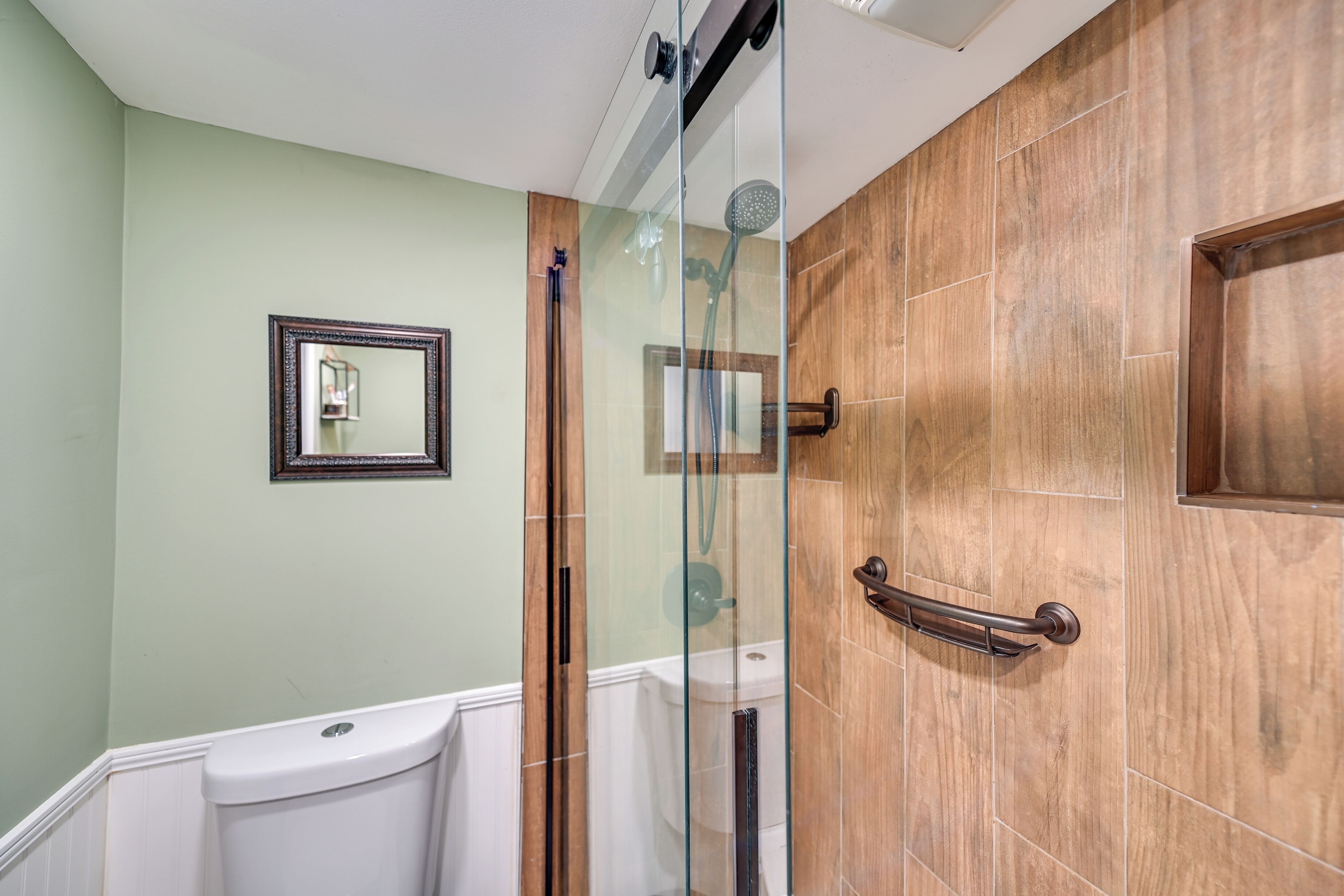 A bathroom with a glass-enclosed shower, a wooden door, a white toilet, and a framed mirror on the wall.