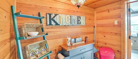 Un intérieur en bois comprenant un espace cuisine, avec le mot « CUISINE » écrit sur le mur, des paniers en osier, un meuble et une poubelle.