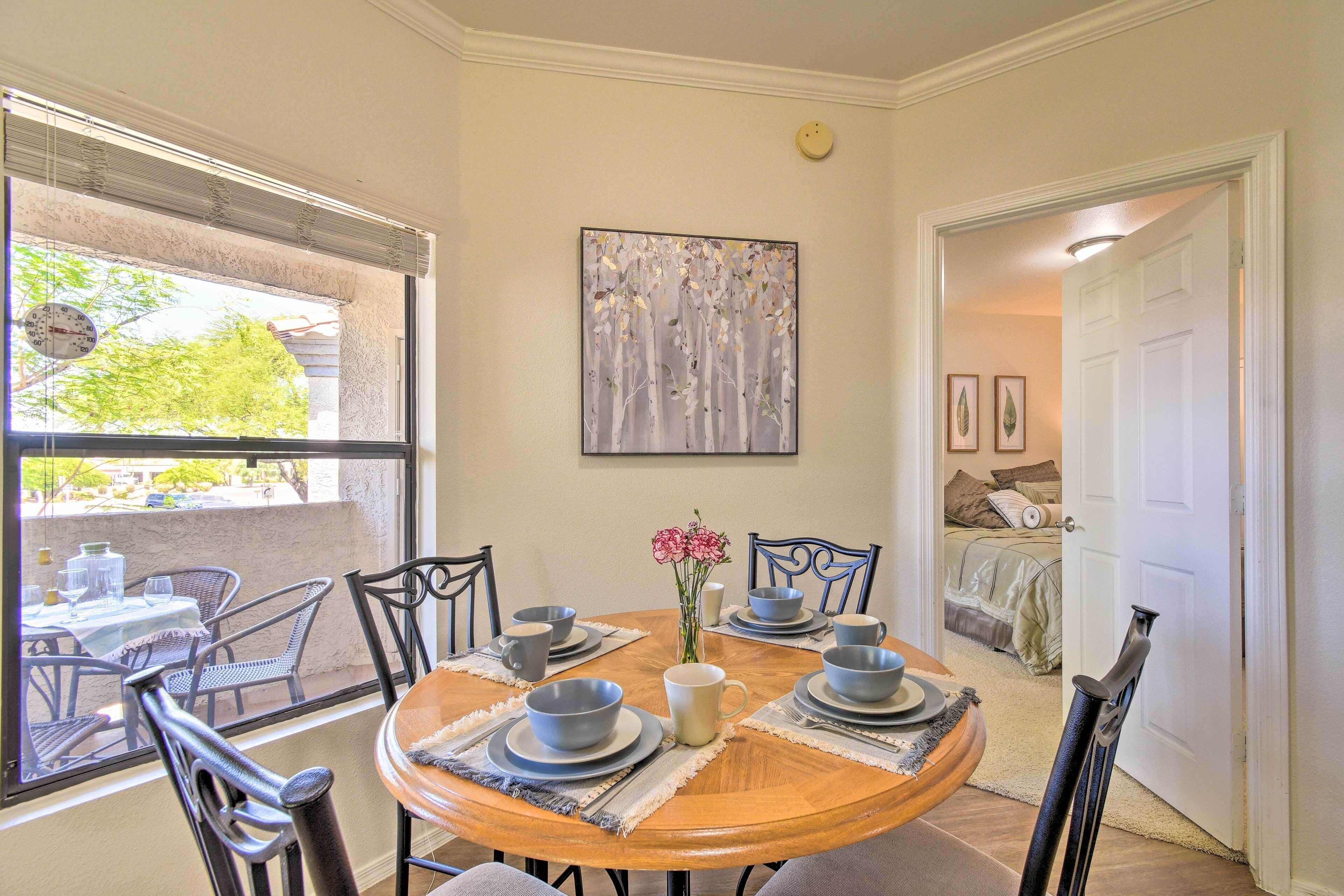 A dining area with a round table set for four, surrounded by chairs, and a window with blinds.