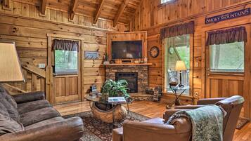 A log cabin interior with a stone fireplace, leather sofas, a television, and a sign reading "Welcome Cabin."