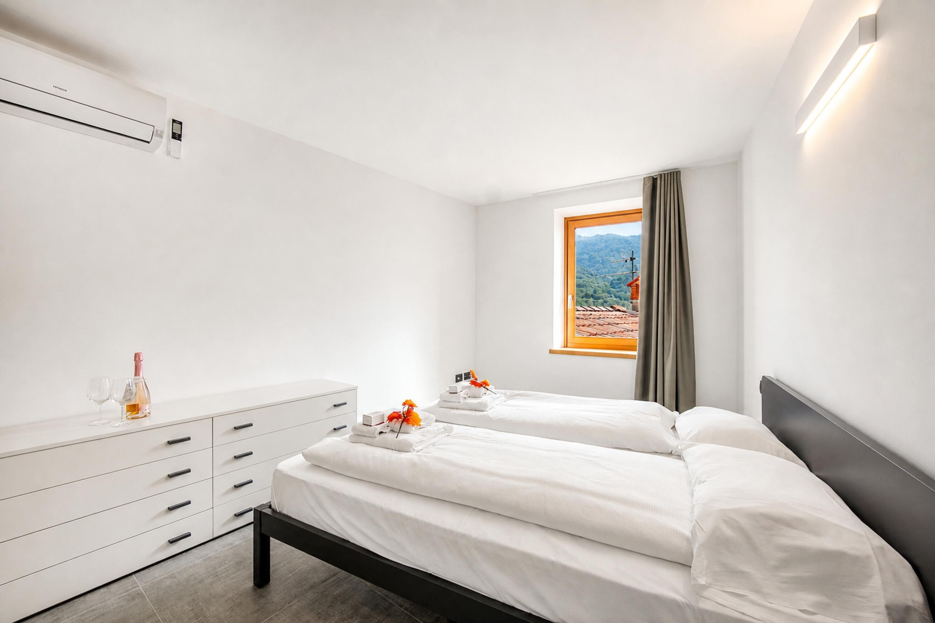 A hotel room with two beds, a white dresser, a window with a view, and a wall-mounted air conditioner.