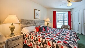 A neatly made bed with a geometric-patterned bedspread, two bedside lamps, a wooden headboard, a framed picture on the wall, a window with red curtains, and a ceiling fan. - Pigeon Retreat w/ Community Pool & Hot Tub! (Pigeon)