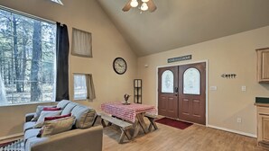 Un salon chaleureux avec un canapé, une table en bois recouverte d’une nappe à carreaux, une horloge et une porte sur laquelle est inscrit le mot « MAISON ».