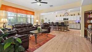 A spacious living room with a leather sofa, a coffee table, and a dining area with chairs. A kitchen with white cabinets and a clock is visible in the background. - Greenview Bliss By Shine Villas #031 5 Bedroom Villa (Davenport)
