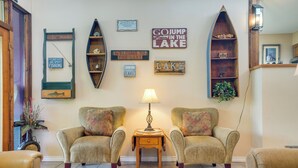 A cozy living room with two armchairs, a wooden coffee table, and wall-mounted shelves with decorative items and signs. - Deck & Gas Grill: Riverfront Mcqueeney Duplex (McQueeney)