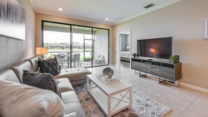 A modern living room with a sofa, coffee table, TV, and a sliding glass door leading to an outdoor area. - Chic Country Club Condo With Pool Access! (Naples)