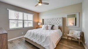 A bedroom with a large bed, a ceiling fan, a window with blinds, a bedside table with a lamp, and a chair with a blanket. - Chic Country Club Condo With Pool Access! (Naples)