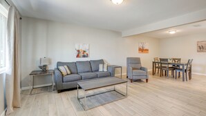 A modern living room with a grey sofa, a coffee table, a side table with a lamp, and a dining area with a table and chairs.